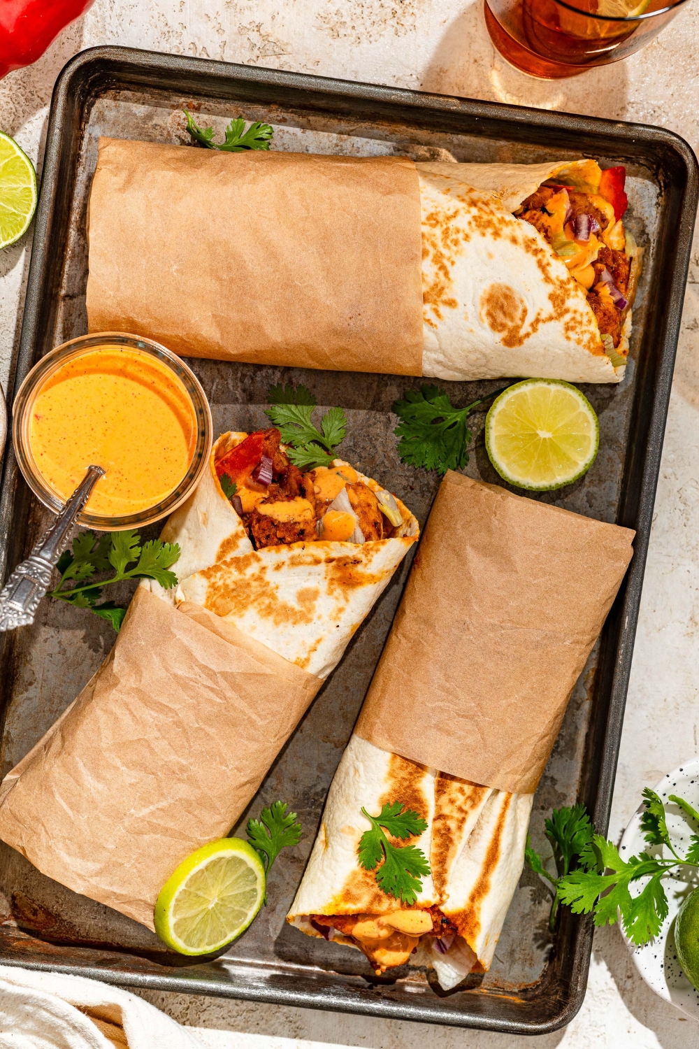 A baking sheet with several honey chipotle chicken wraps wrapped in paper and garnished with cilantro and lime. The wraps are served with a bowl of dipping sauce. The sheet is on a tan counter with garnishes and a bowl of salt.