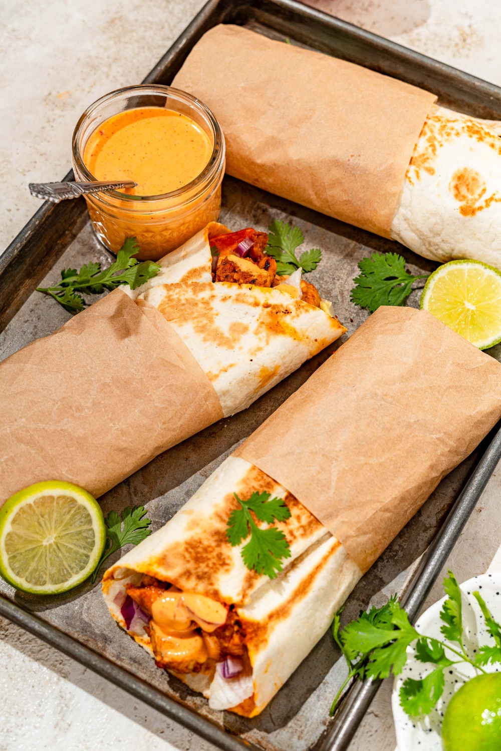 A baking sheet with several honey chipotle chicken wraps wrapped in paper and garnished with cilantro and lime. The wraps are served with a bowl of dipping sauce. The sheet is on a tan counter with garnishes and a bowl of salt.