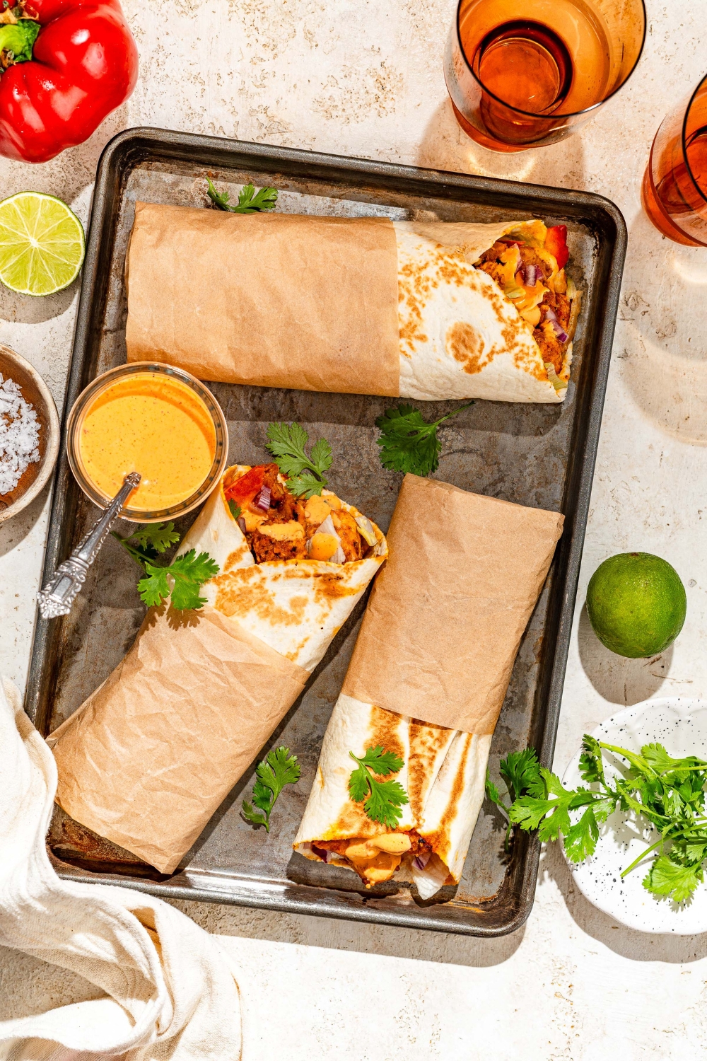 A baking sheet with several honey chipotle chicken wraps wrapped in paper and garnished with cilantro and lime. The wraps are served with a bowl of dipping sauce. The sheet is on a tan counter with garnishes and a bowl of salt.