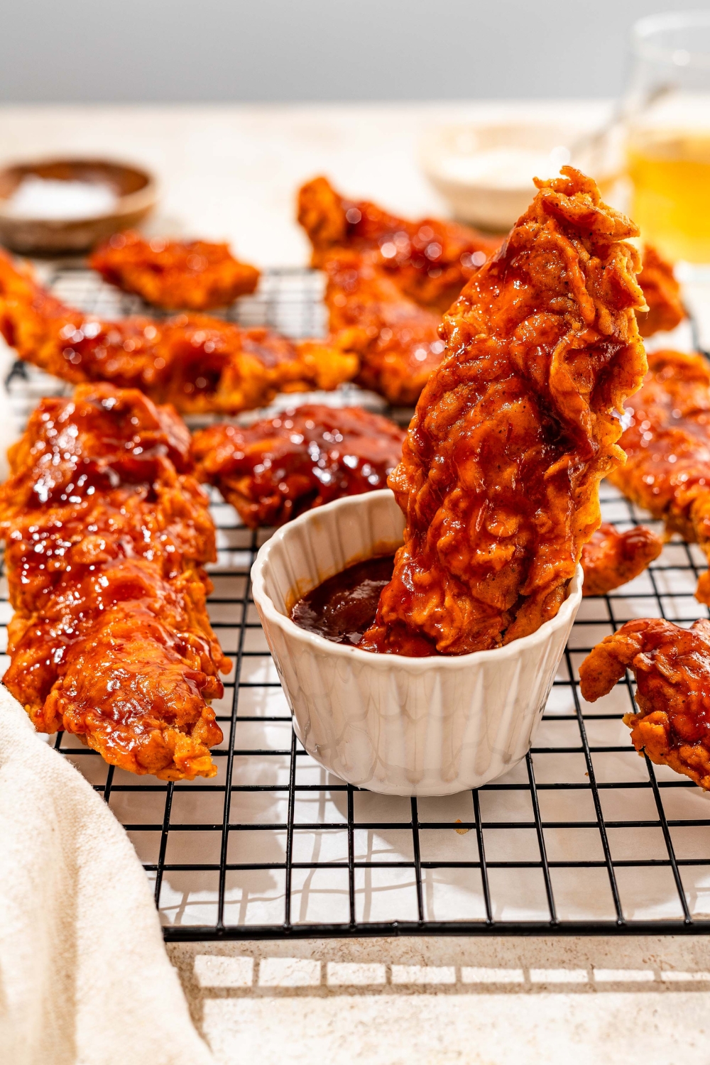 A wire rack with honey BBQ chicken tenders and a bowl of honey BBQ sauce. There is a piece of chicken dipped in the sauce. The rack is on a tan counter.