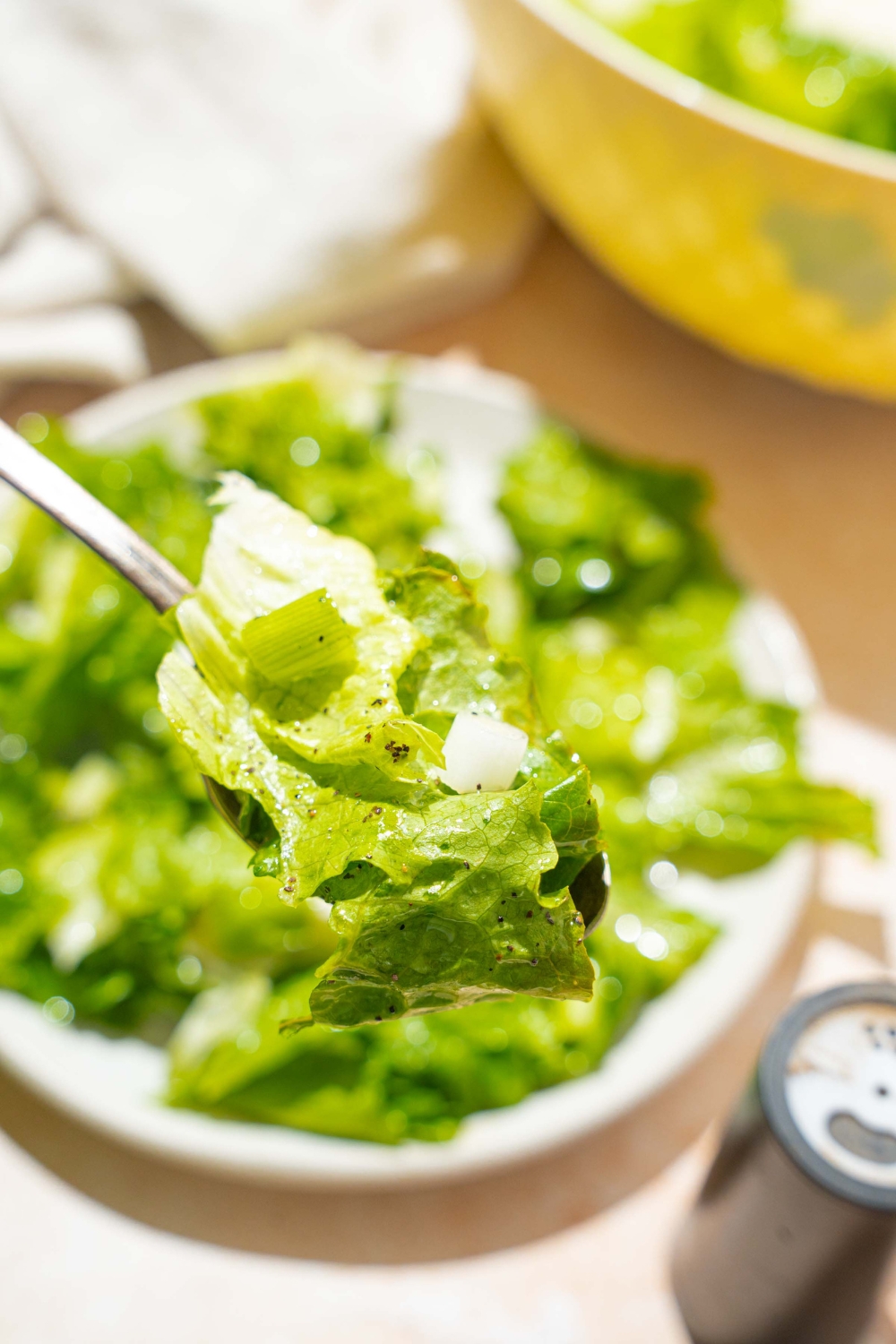 A close up of a fork with a bite of wilted lettuce salad tossed in a bacon vinegar dressing. There is a bowl of salad on a tan counter blurred in the background.