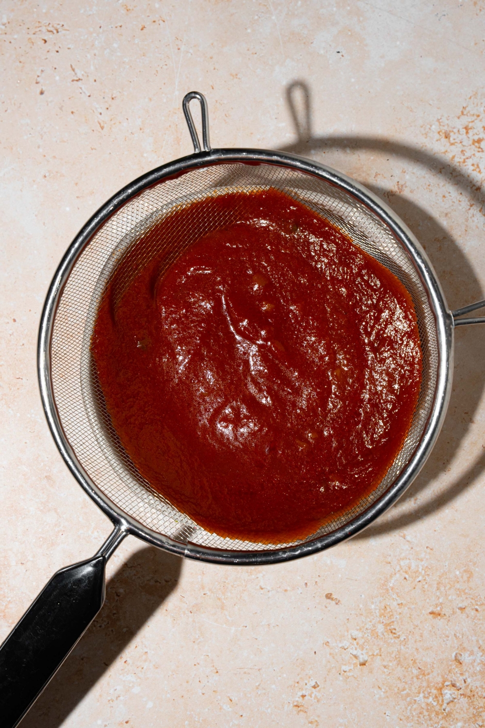 A copycat Bachan Japanese BBQ sauce mixture in a strainer over a bowl. The bowl is on a tan counter.