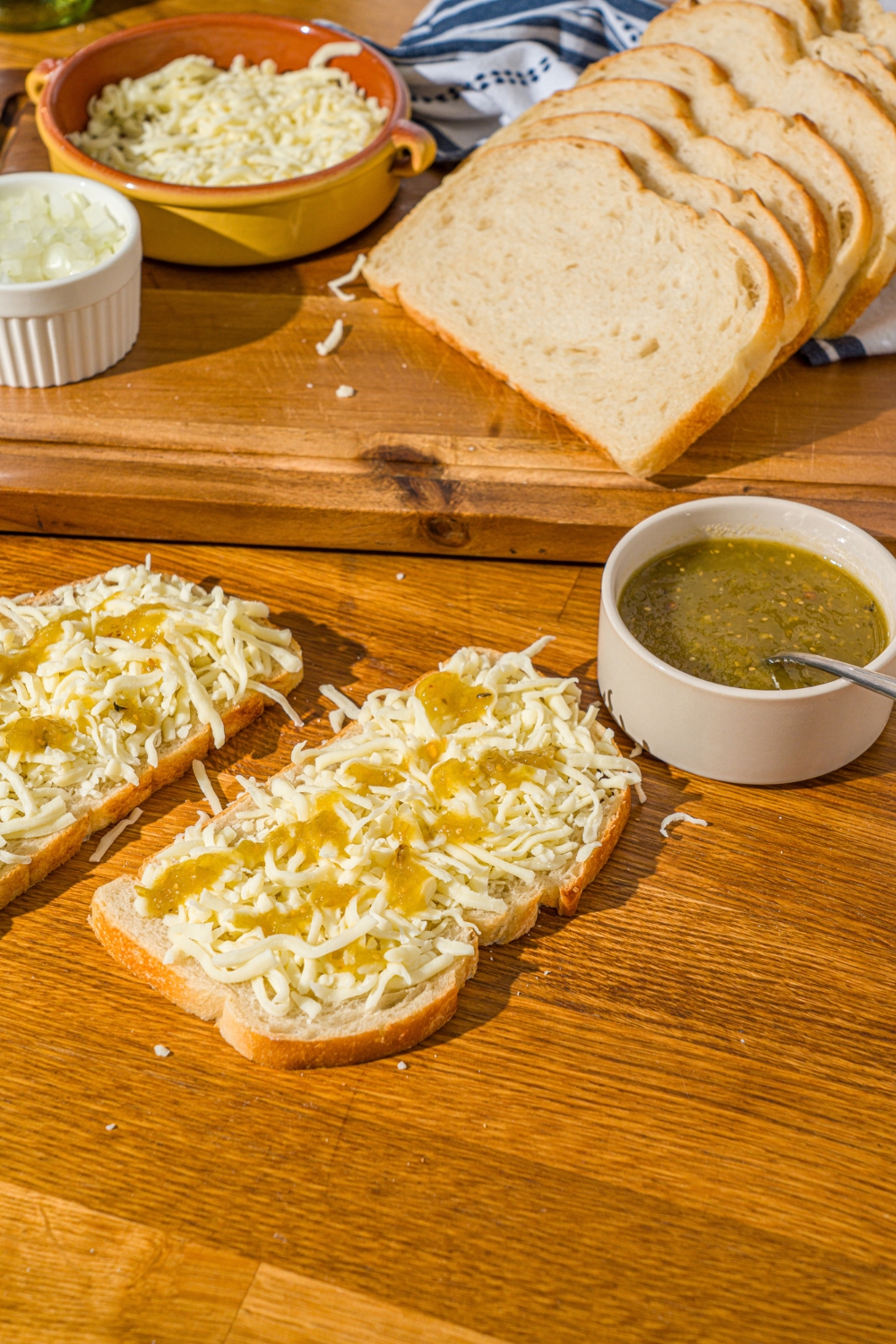 Two slices of sourdough bread with shredded mozzarella cheese drizzled with salsa verde. The bread is on a wooden counter with a bowl of salsa verde and cutting board with ingrediens.