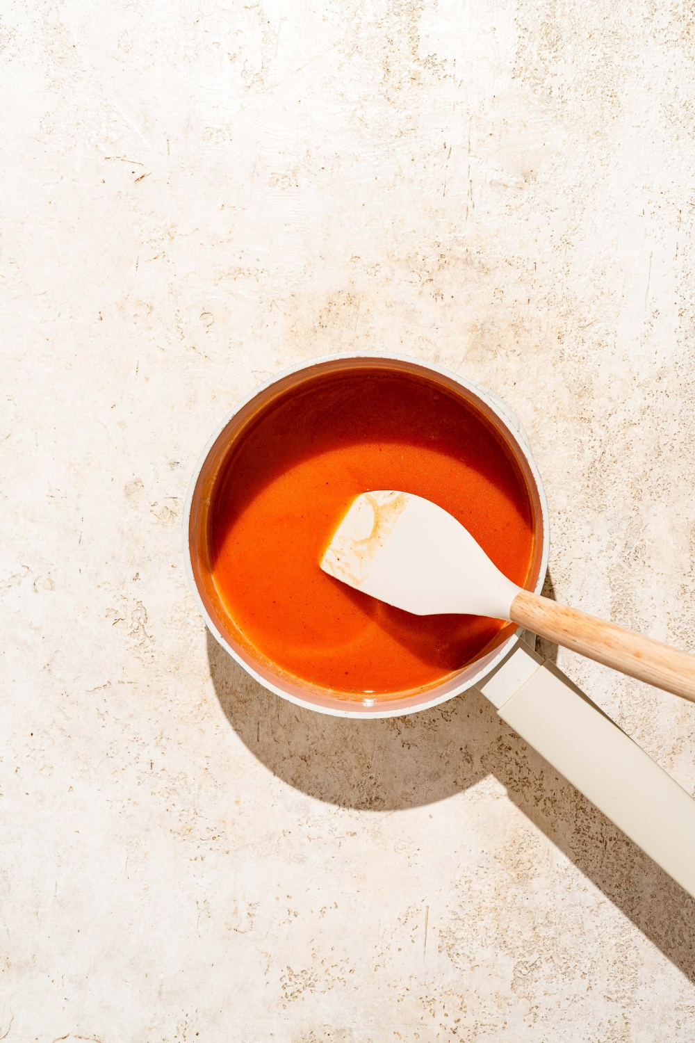 A small saucepan with a spatula stirring Buffalo sauce. The saucepan is on a tan counter.
