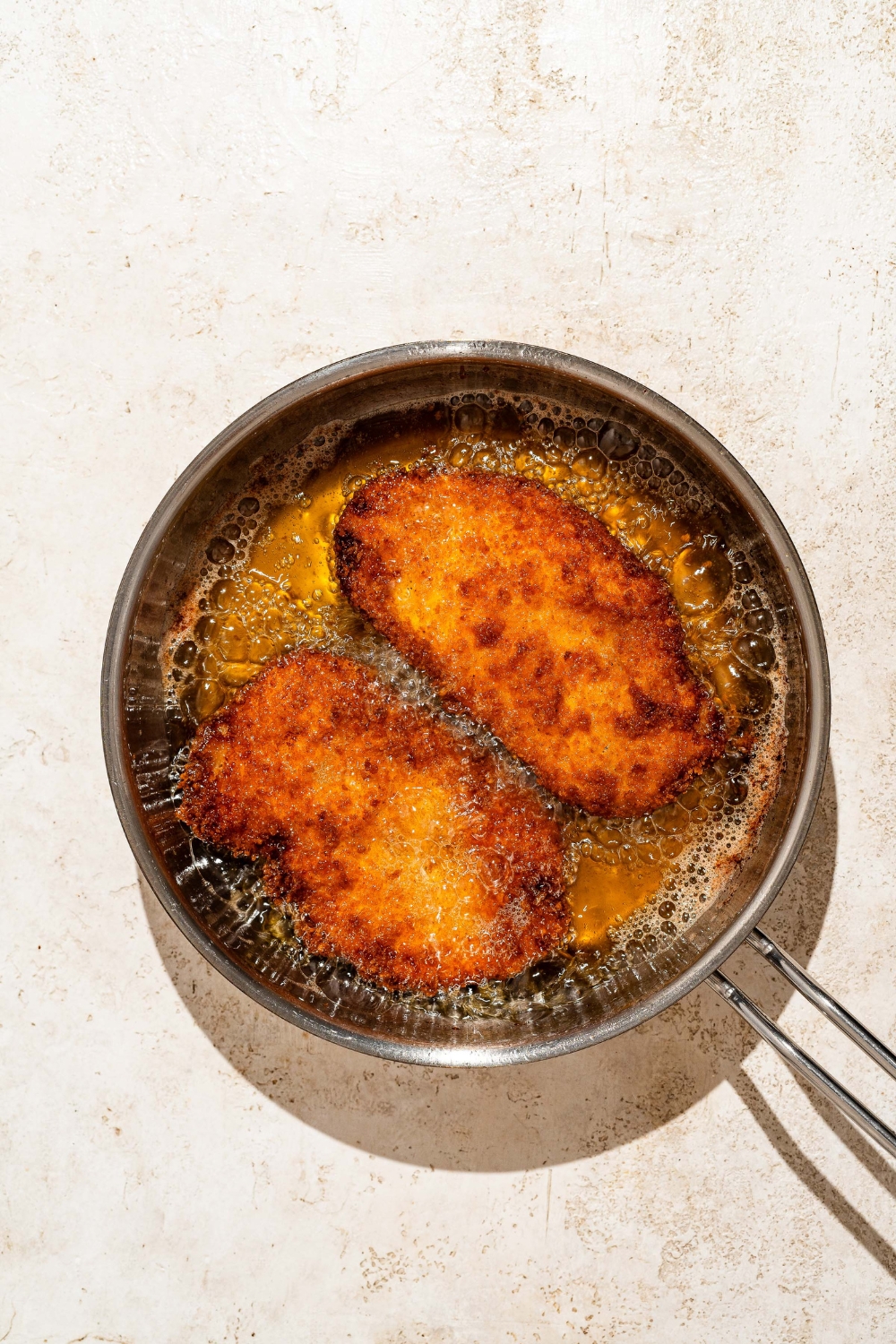 A frying pan with two breaded chicken cutlets frying in oil. The pan is on a tan counter.