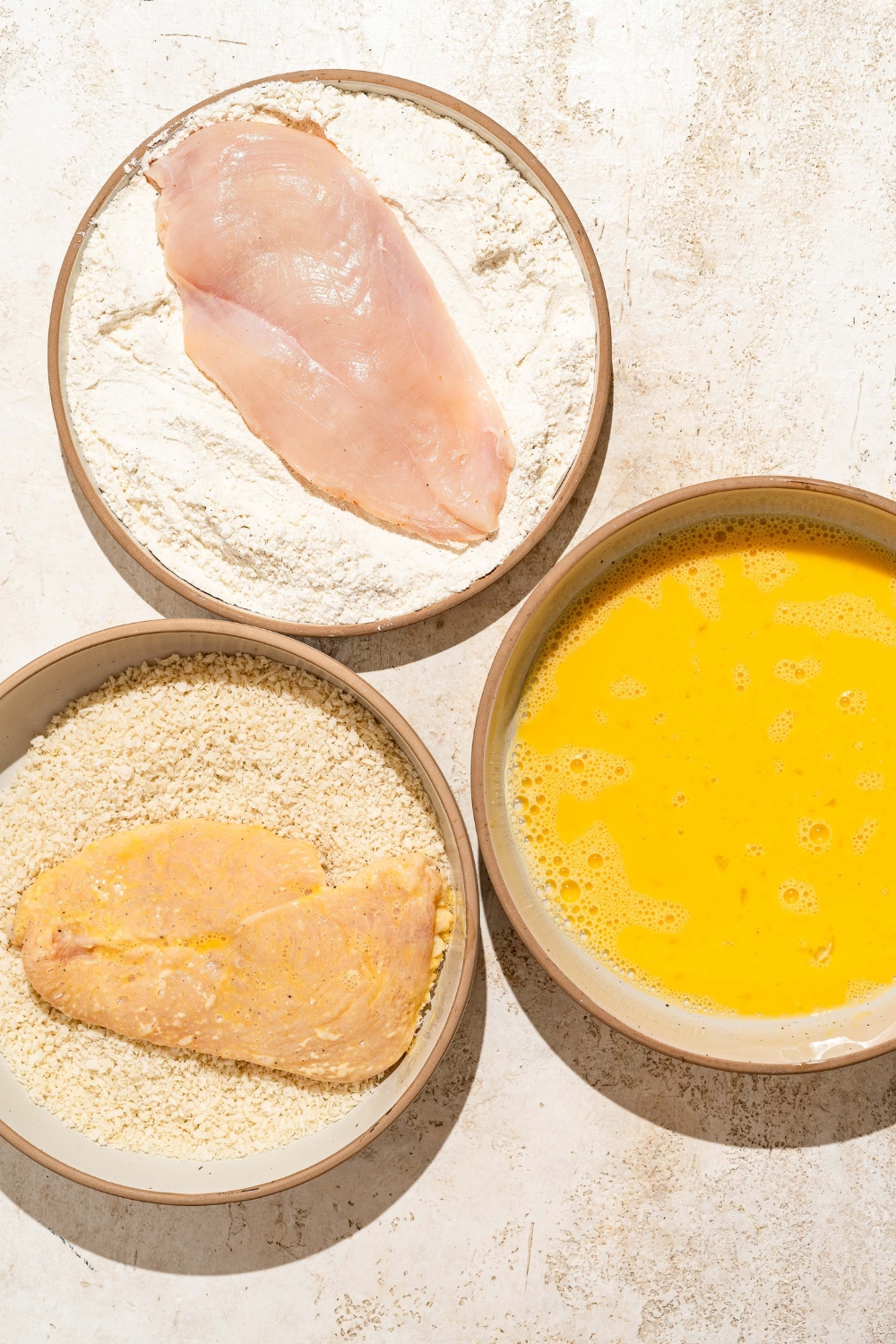 Three plates with ingredients to bread chicken cutlets including whisked egg, flour, and breadcrumbs. There is a chicken cutlet in the flour and a chicken cutlet dipped in egg in the breadcrumbs.