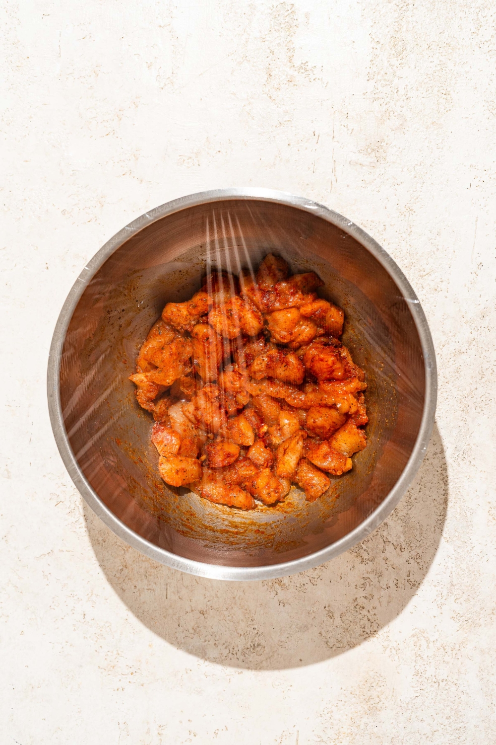 A large bowl with uncooked seasoned Buffalo chicken covered in plastic wrap. The bowl is on a tan counter.