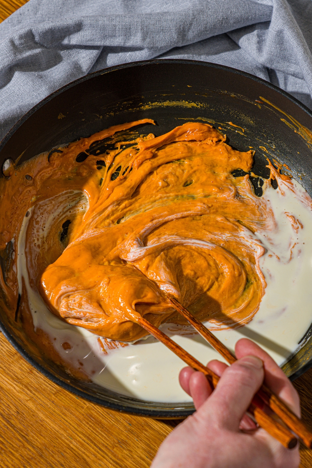 A skillet with a pair of chopsticks stirring melted cheese, Buldak sauce, egg yolks, and heavy cream. The skillet is on a wooden counter with a blue cloth napkin.