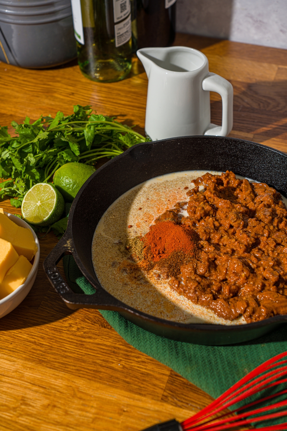 A skillet with ingredients to make Chili's skillet queso including Hormel chili, milk, and seasonings. The skillet is on a wooden counter with a green cloth napkin, cilantro, and lime.