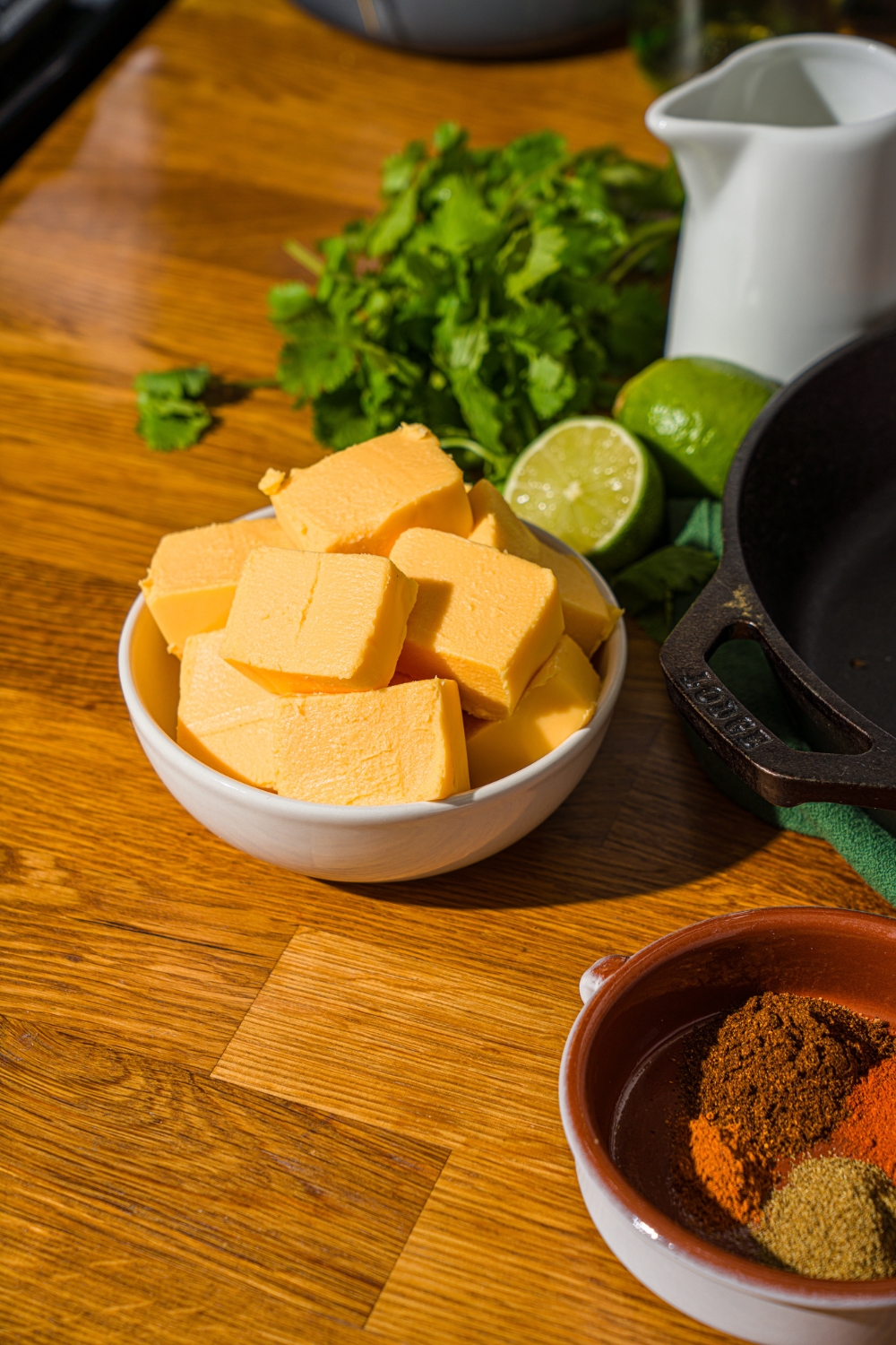 A small white bowl with cubed Velveeta cheese. The bowl is on a wooden counter with a bowl of seasonings, cilantro, and lime.