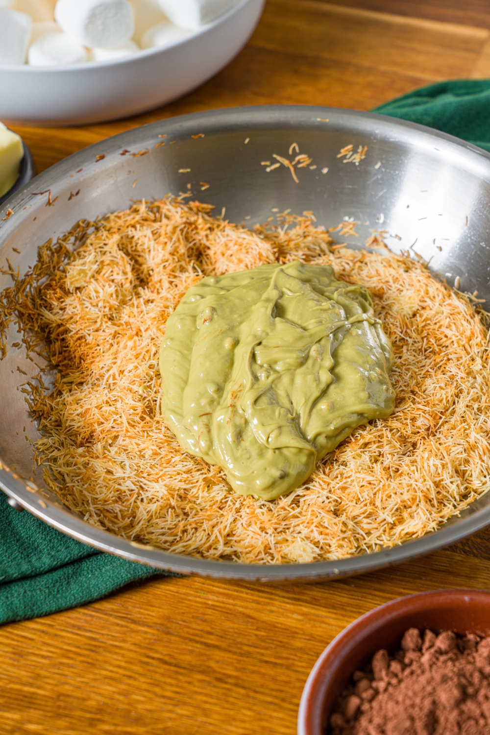A large bowl with toasted katafi topped with pistachio cream. The bowl is on a wooden counter with a green cloth napkin and bowl of cocoa powder.