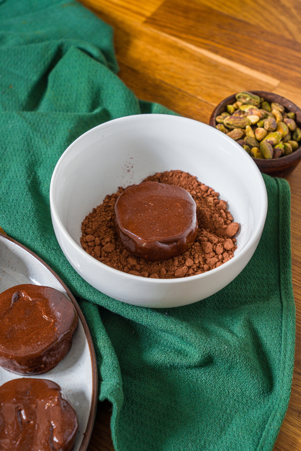 A Dubai chocolate cookie dipped in a bowl of cocoa powder. The bowl is on a wooden counter with a green cloth napkin.