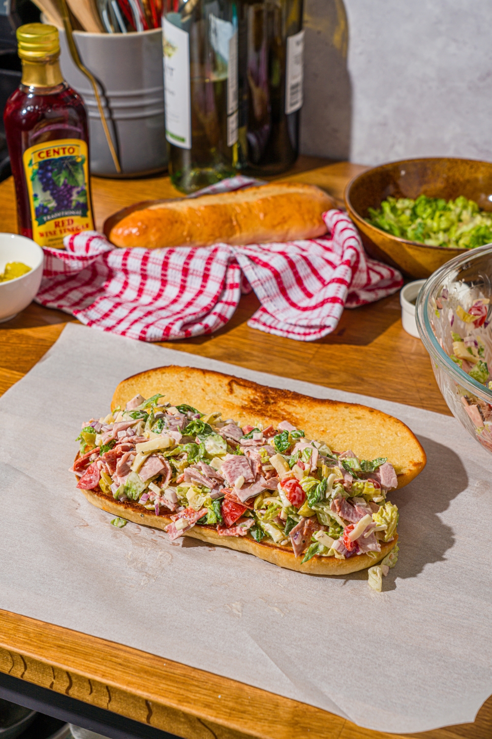 A piece of parchment paper with a whole open chopped Italian sandwich. The sandwich is on a wooden counter with a red striped napkin.