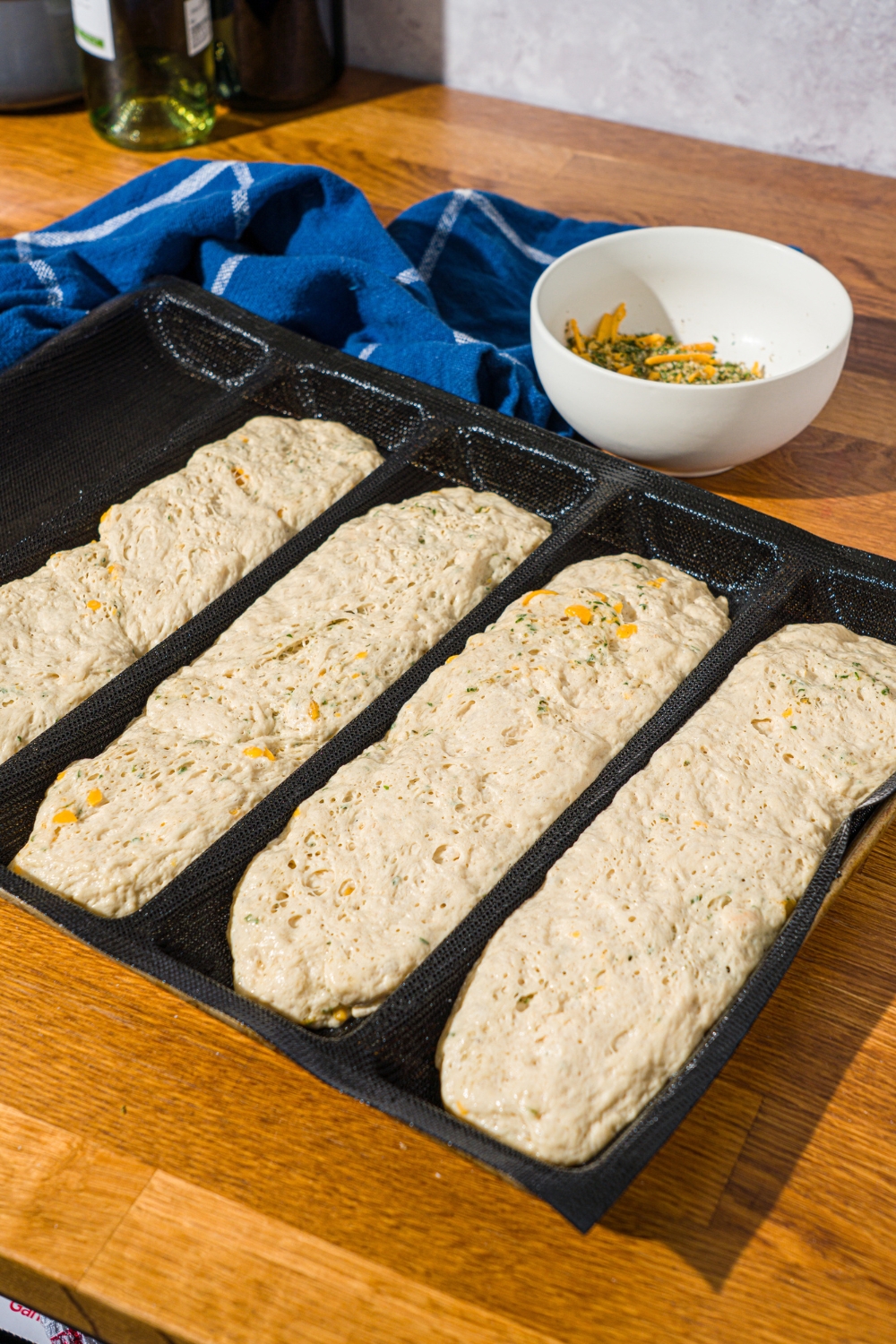 Several Italian herb and cheese bread dough loaves proofing in a loaf pan. The pan is on a wooden counter with a blue striped napkin.