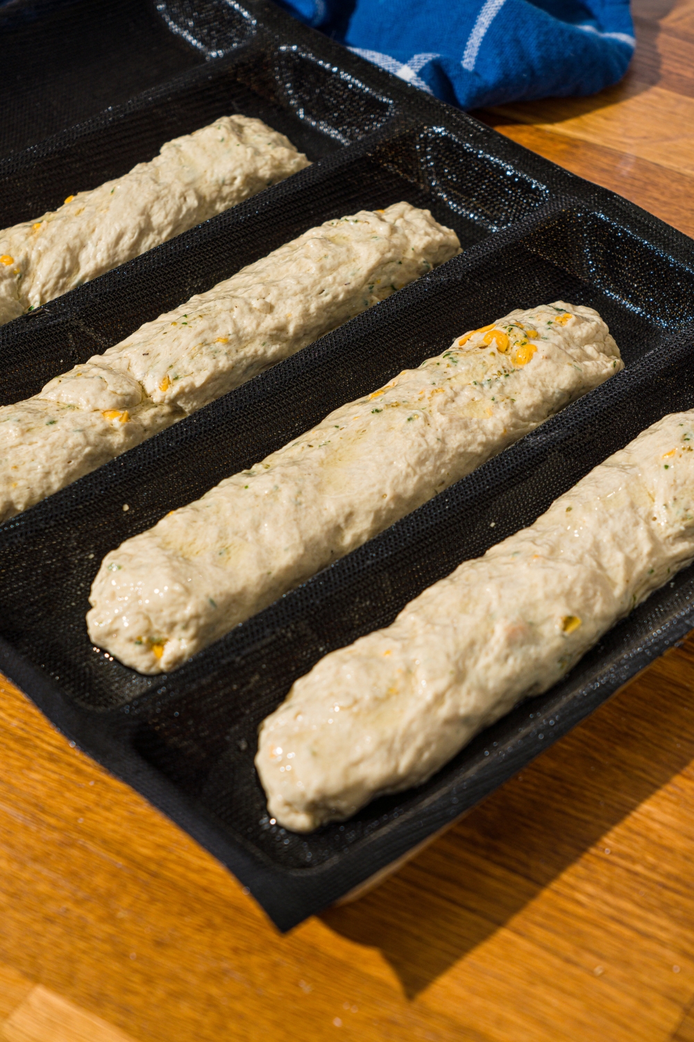 Several Italian herb and cheese bread dough loaves proofing in a loaf pan. The pan is on a wooden counter with a blue striped napkin.
