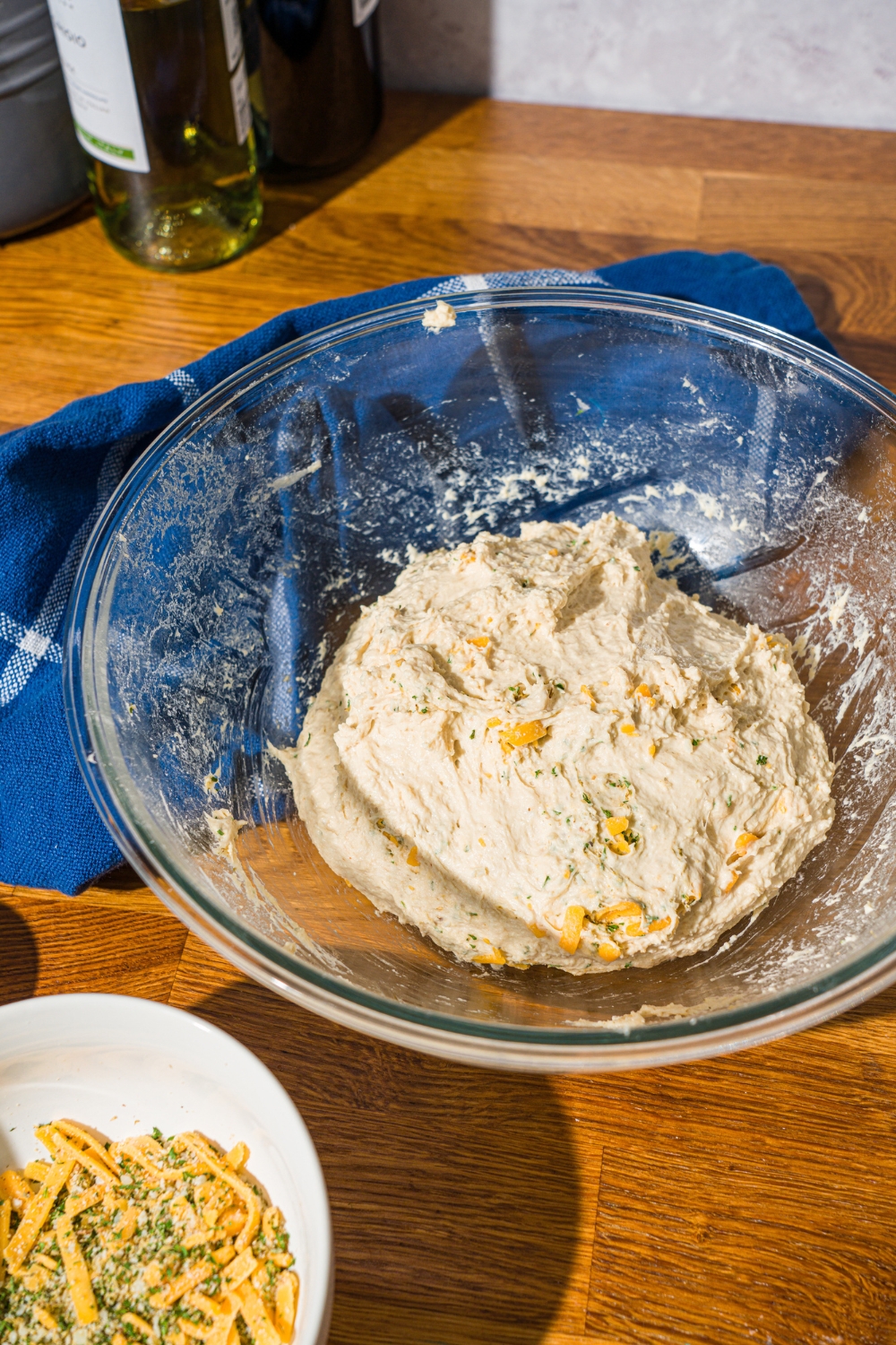 A glass bowl with kneaded Italian herb and cheese bread dough mixed with shredded cheese and herbs. The bowl is on a wooden counter with a blue cloth napkin.