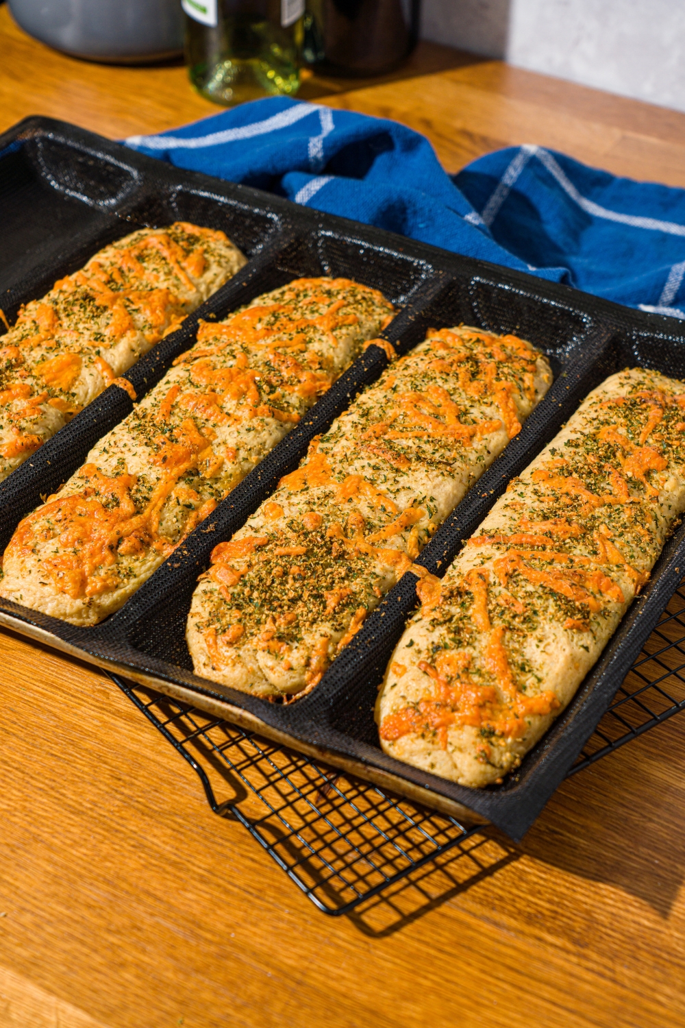 Several baked Italian herb and cheese bread loaves in a loaf pan. The pan is on a wire rack on a wooden counter with a blue cloth napkin.
