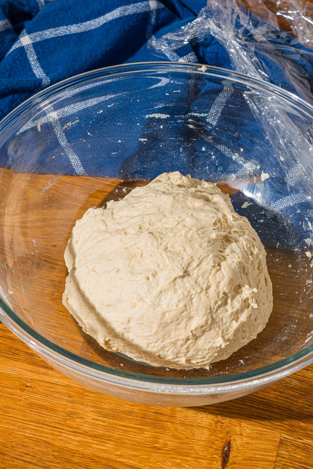 A glass bowl with a kneaded bread dough ball. The bowl is on a wooden counter with a blue cloth napkin.