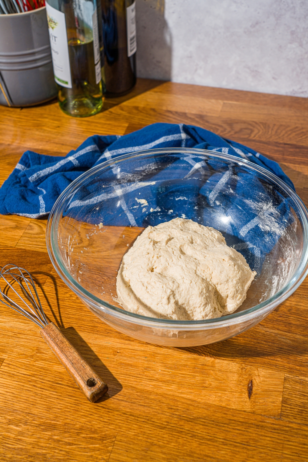 A glass bowl with kneaded bread dough on a wooden counter with a blue cloth napkin.