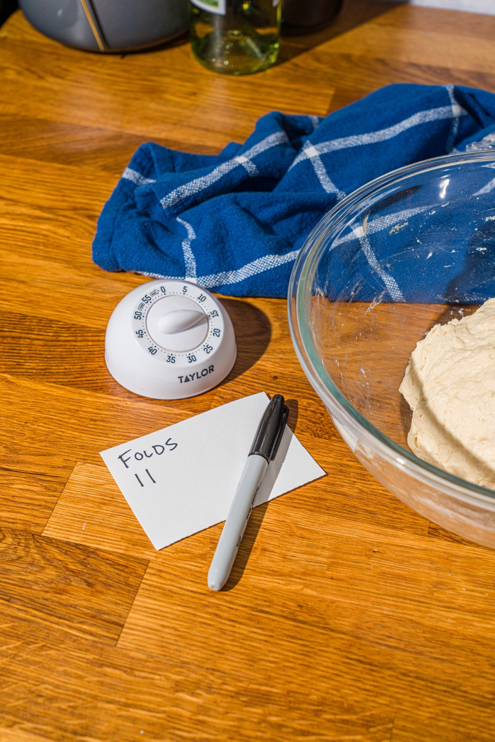 A glass bowl with kneaded bread dough. The bowl is on a wooden counter with a kitchen timer and piece of paper with a sharpie.