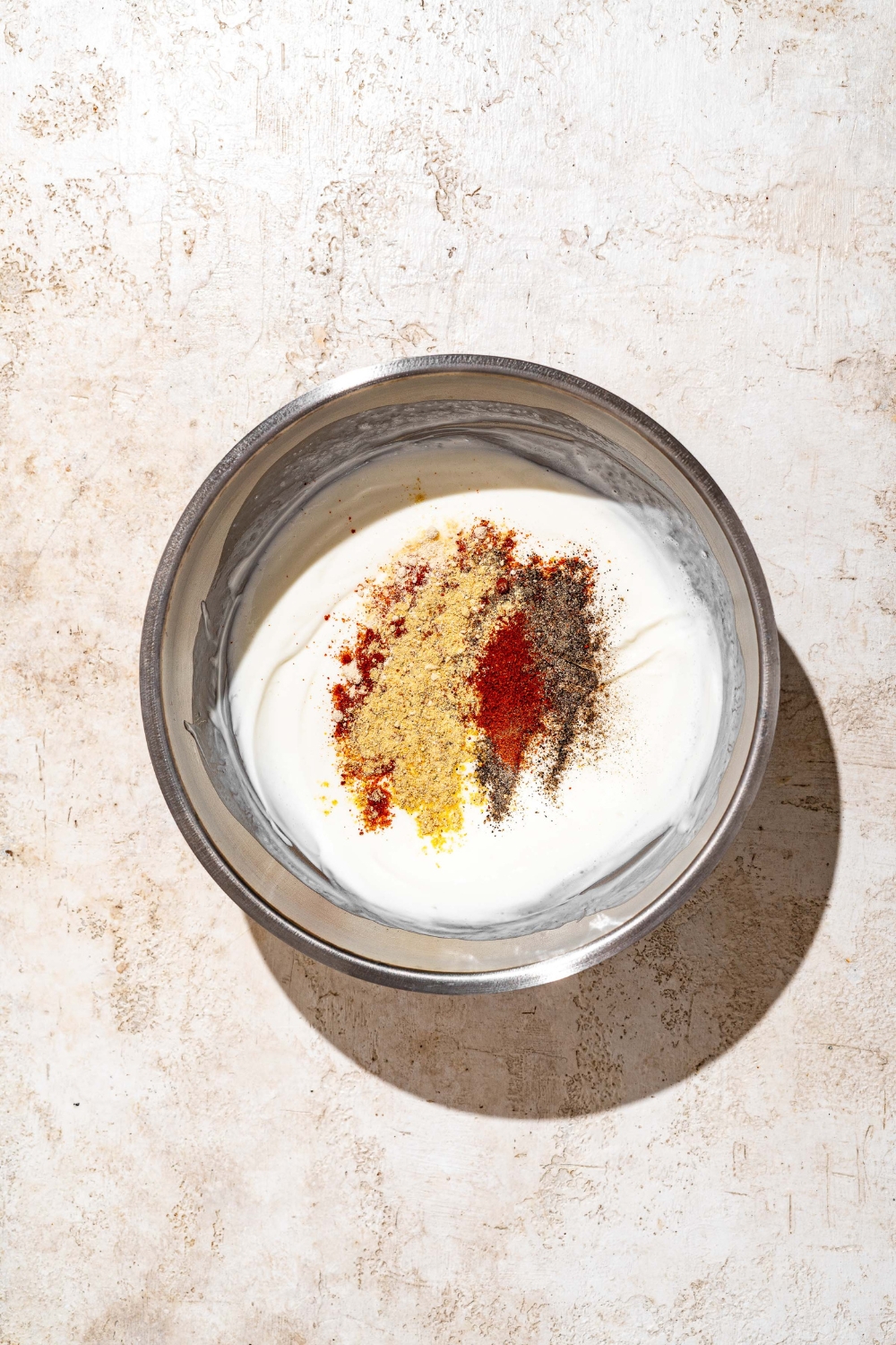 A large bowl with a mayo and buttermilk mixture topped with seasonings. The bowl is on a tan counter.