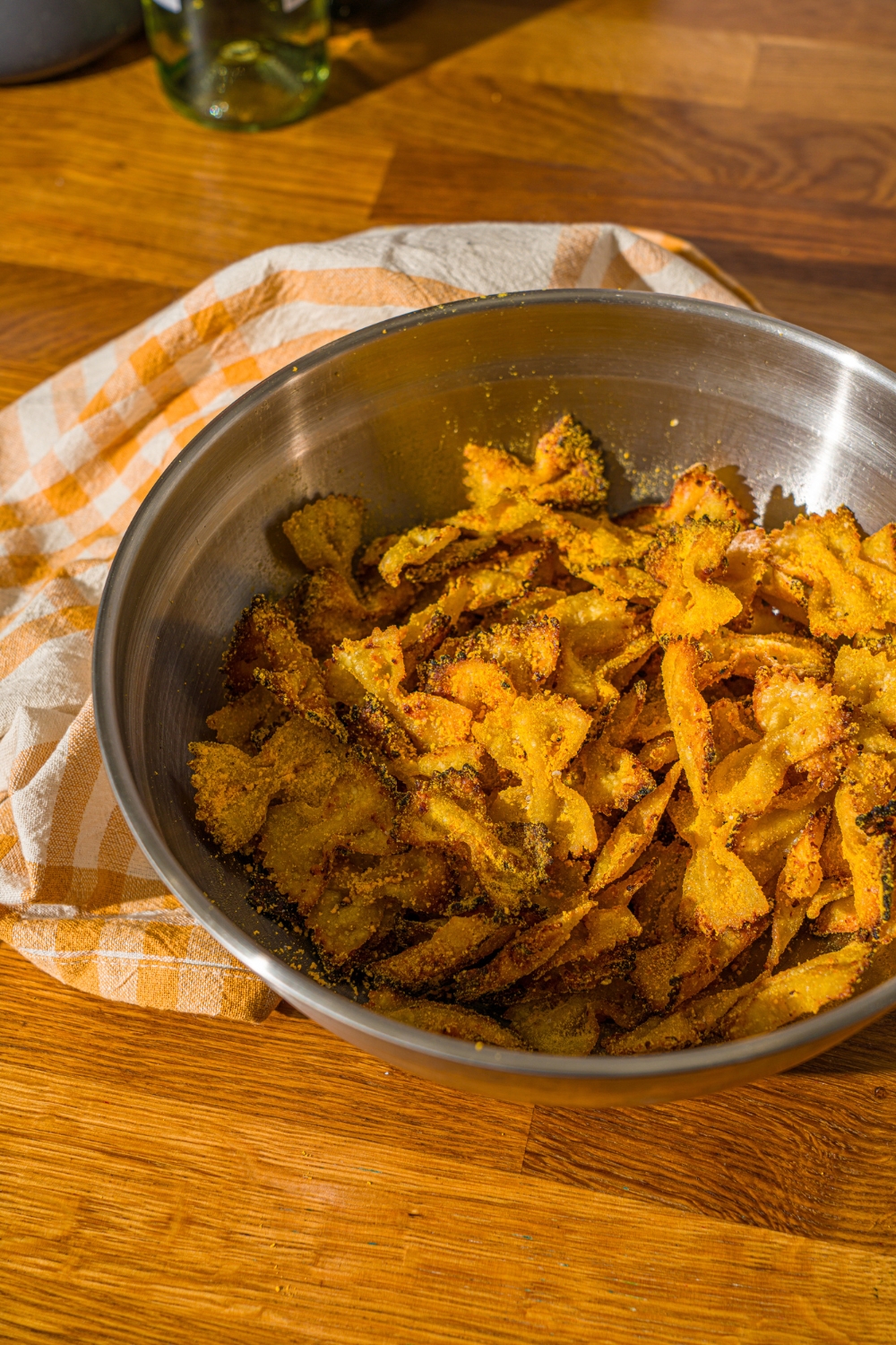 A mixing bowl with air fried bowtie pasta chips tossed in cheese and seasonings. The bowl is on a wooden counter with a yellow striped napkin.
