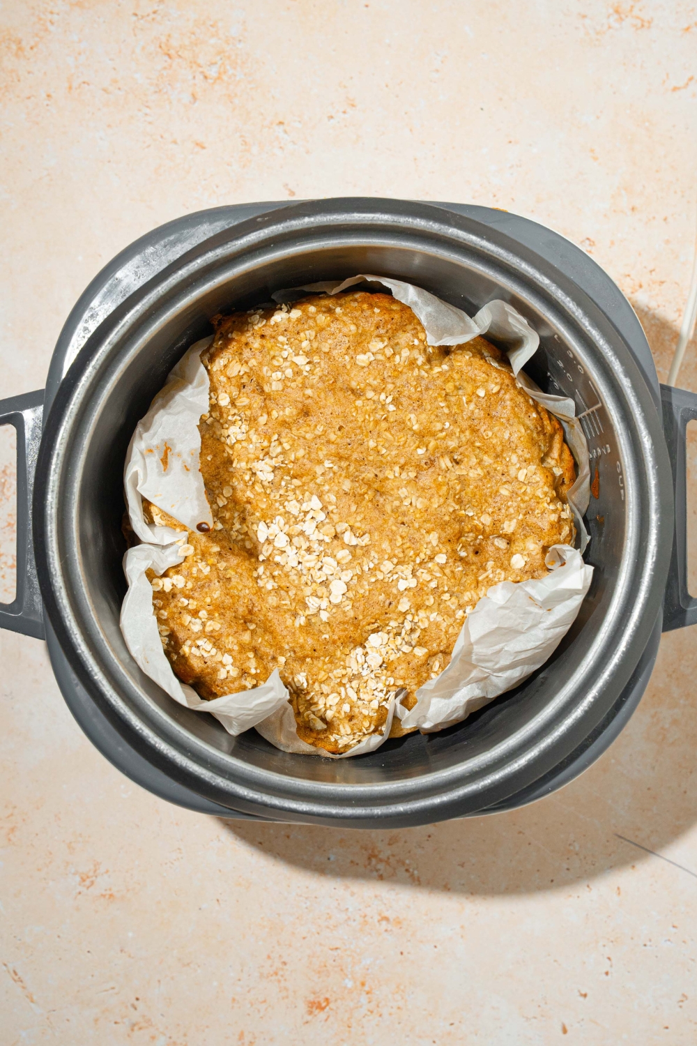 A cooked loaf of rice cooker, banana bread on parchment paper inside of a rice cooker.