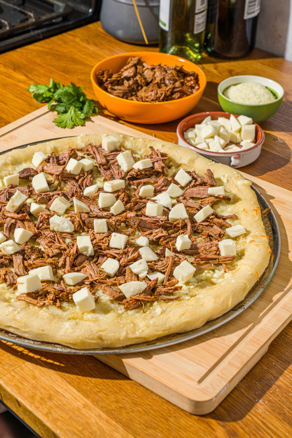 An unbaked birria pizza topped with birria meat and fresh mozzarella on a pizza tray. The tray is on a wooden board.