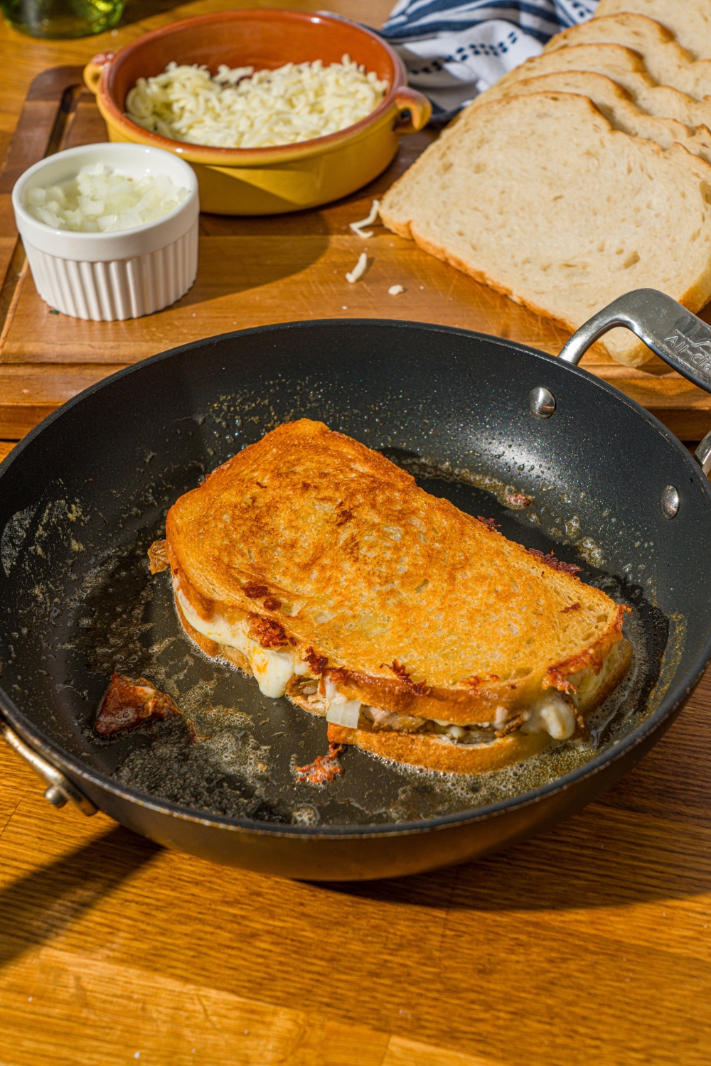 A skillet with a whole birria grilled cheese sandwich cooking in butter. The skillet is on a wooden counter with a cutting board of ingredients.