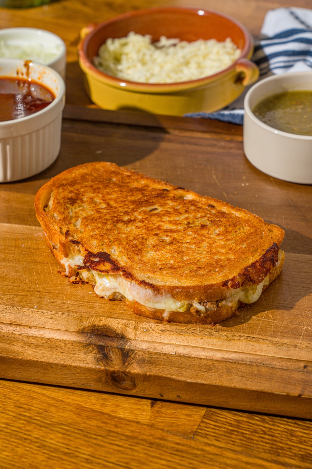 A birria grilled cheese sandwich on a cutting board with bowls of birria sauce and salsa verde. The cutting board is on a wooden counter.