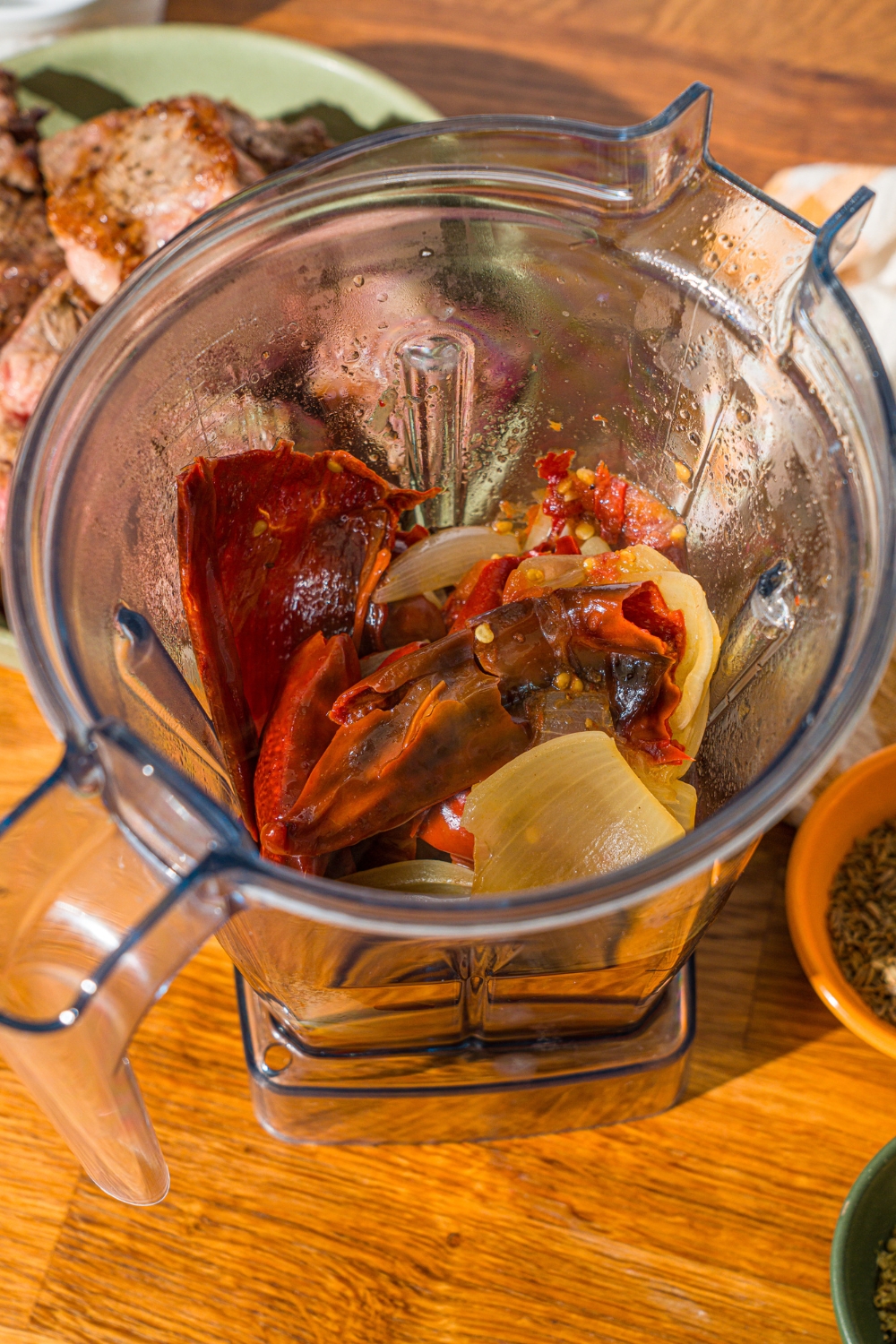 A blender with simmered peppers, onion, tomatoes, and garlic. The blender is on a wooden counter.