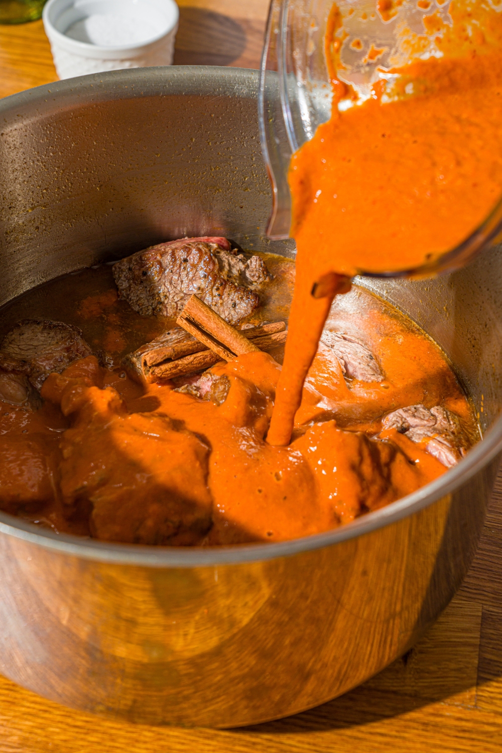 A large stock pot with seared chuck roast in broth and a blender adding pepper sauce. The pot is on a wooden counter.