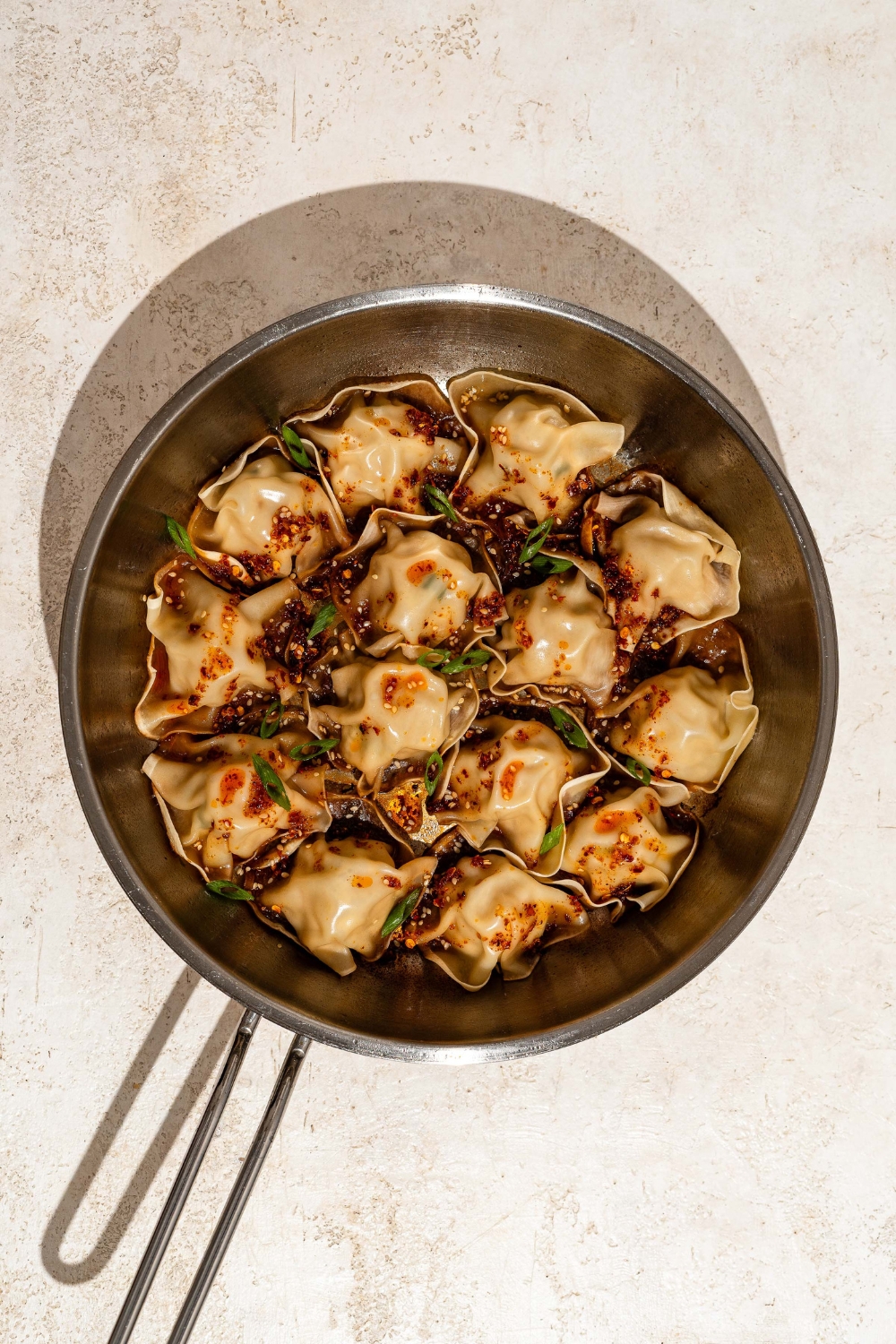 A skillet with blanket dumplings topped with chili crisp, sesame seeds, and sliced green onion. The skillet is on a tan counter.