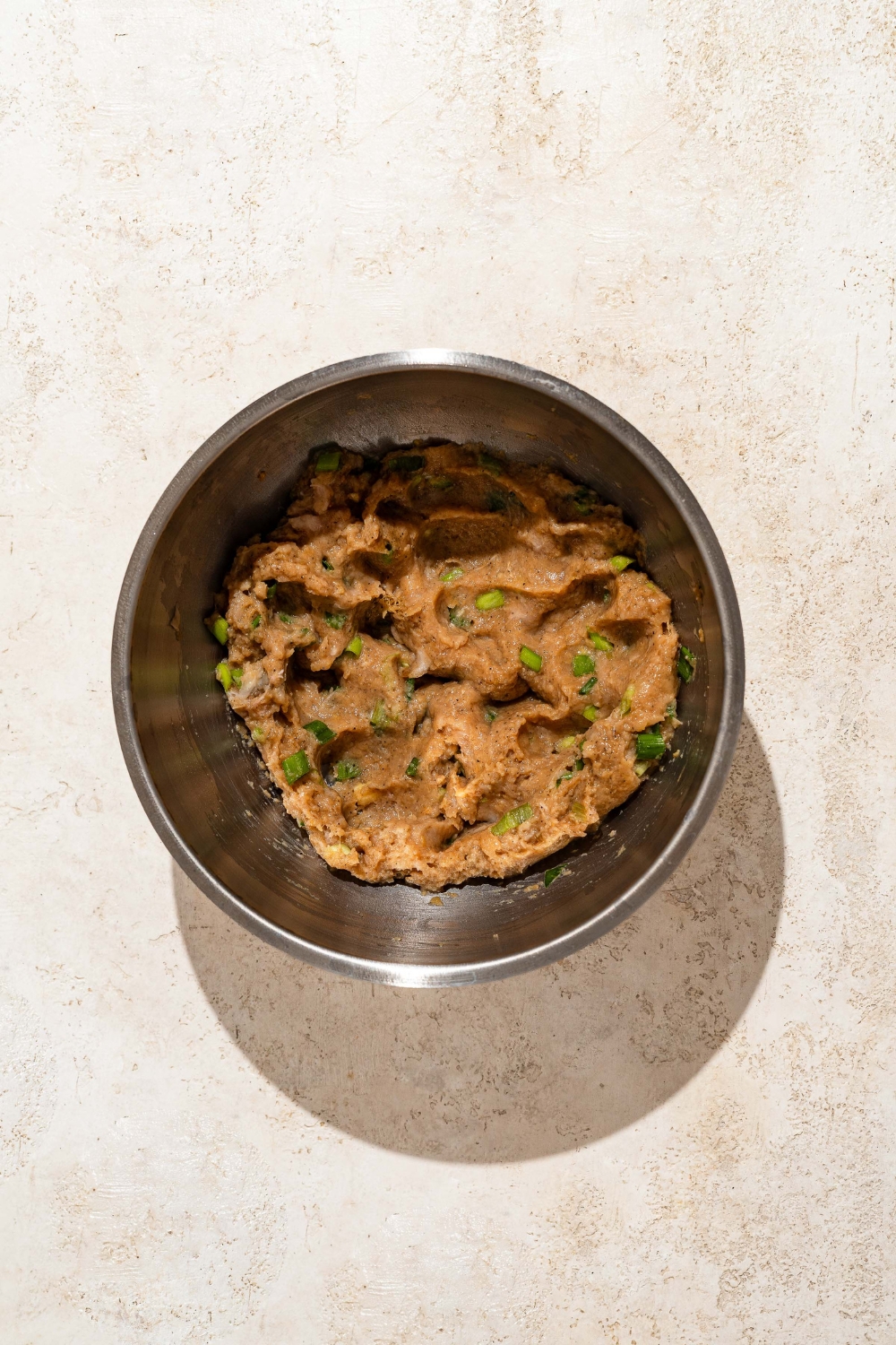 A large bowl with a ground pork mixture including garlic, green onion, soy sauce, ginger paste, sesame oil, and more. The bowl is on a tan counter.
