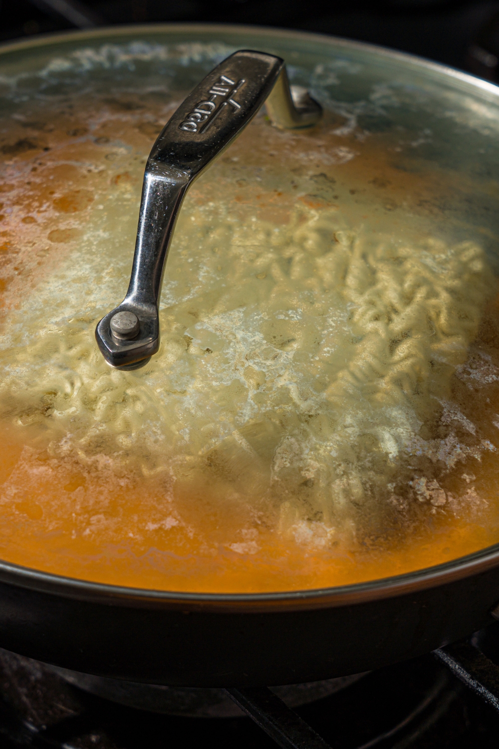 A covered skillet with Buldak ramen noodles on cheese sauce. The skillet is cooking on the stove.