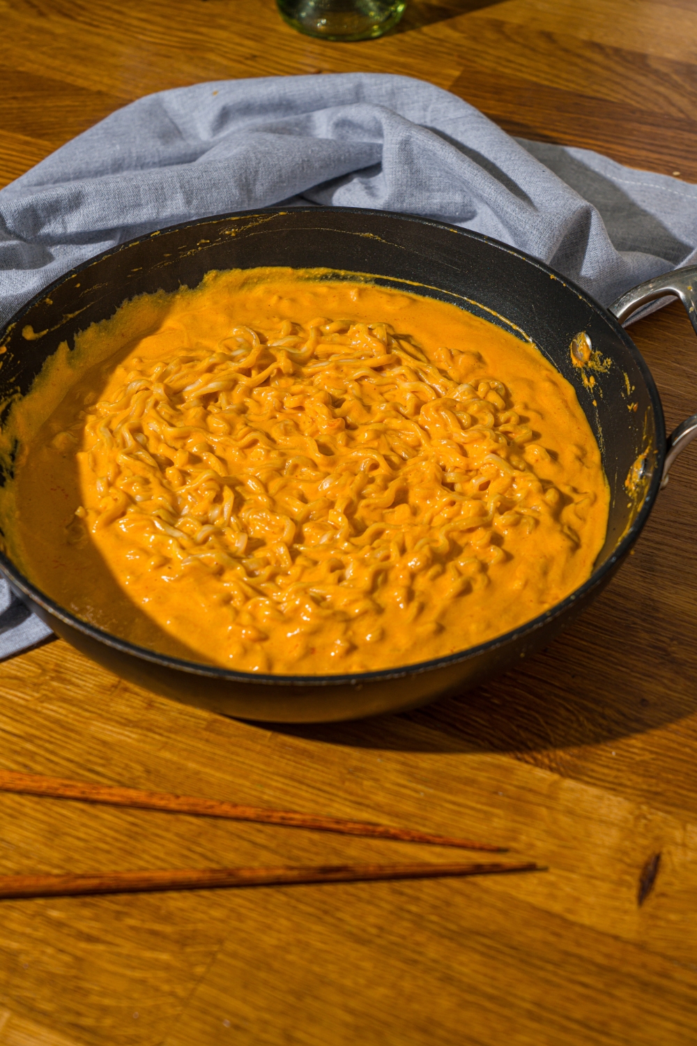A skillet with cooked ramen noodles in a Buldak ramen sauce. The skillet is on a wooden counter with a blue cloth napkin and pair of chopsticks.