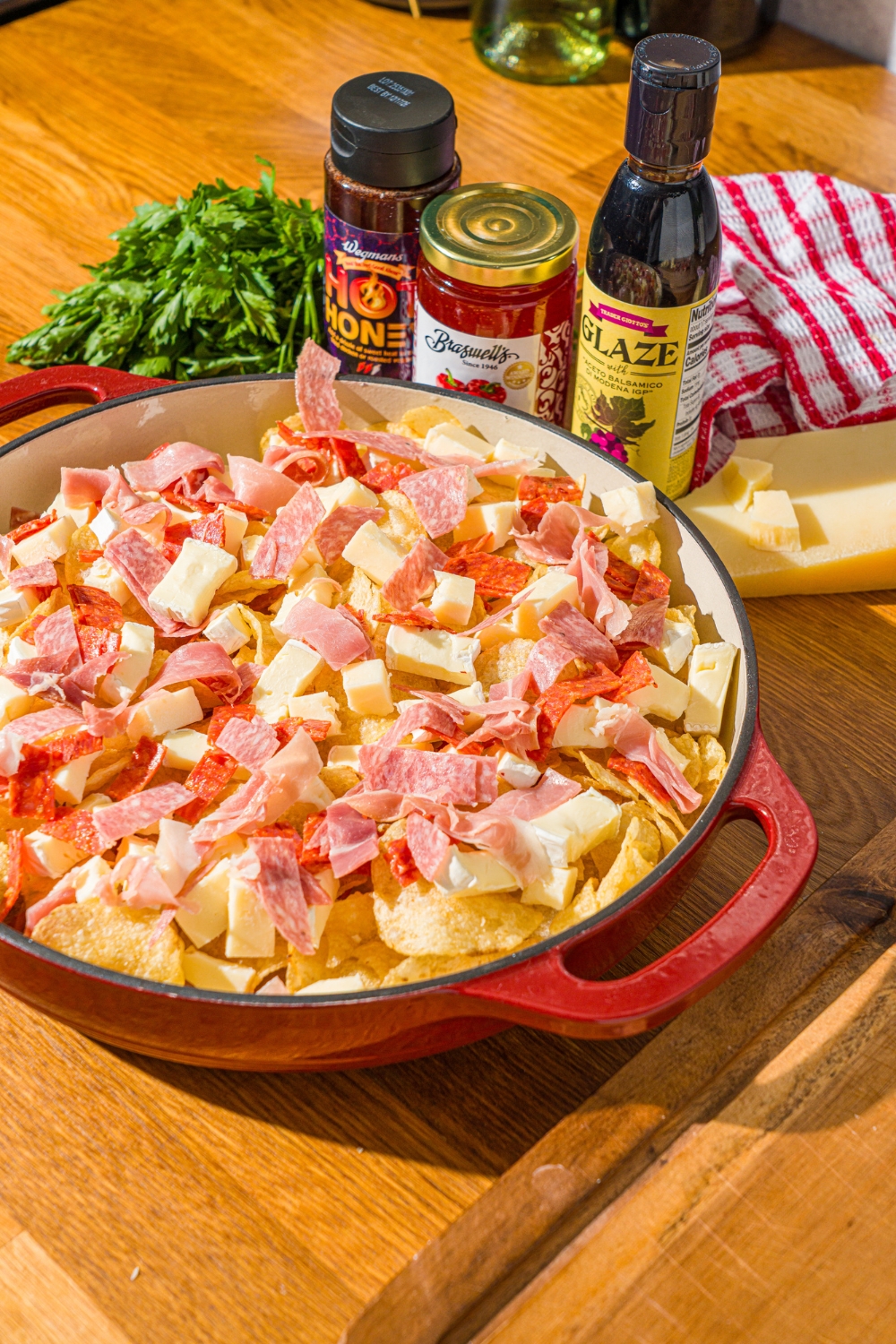 A baking dish with kettle chips layered with charcuterie meats and cheeses like brie, cheddar cheese, prosciutto, salami, and pepperoni. The dish is on a wooden board with jars of toppings.