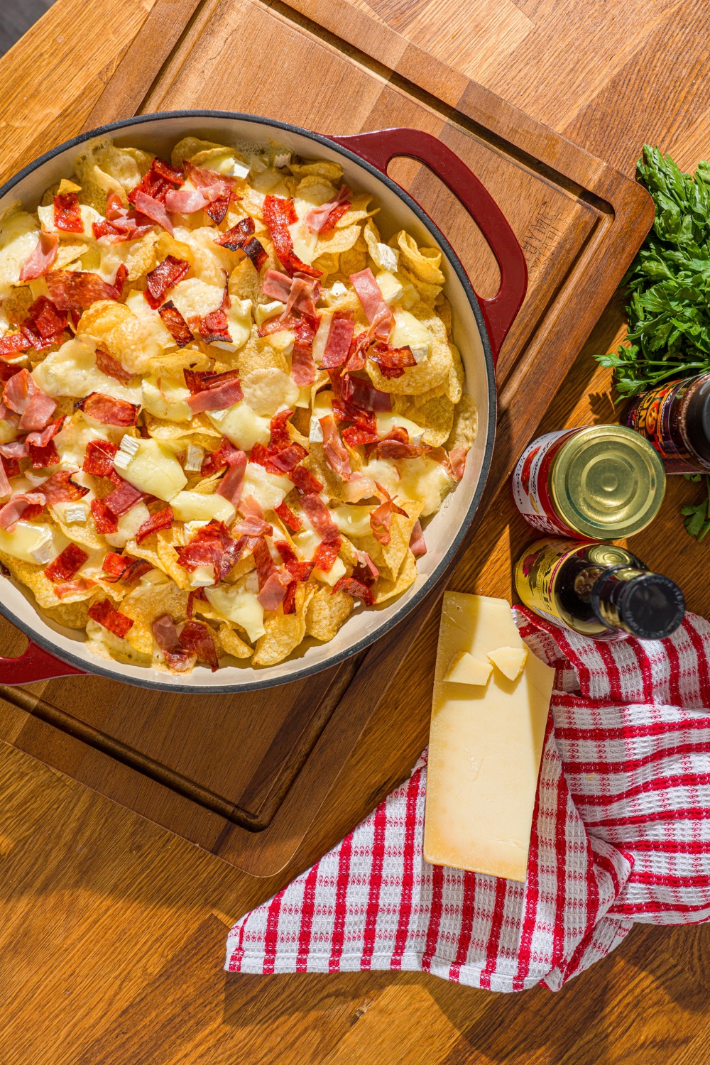 A baking dish with baked charcuterie nachos. The dish is on a wooden board with parmesan cheese and jars of toppings.
