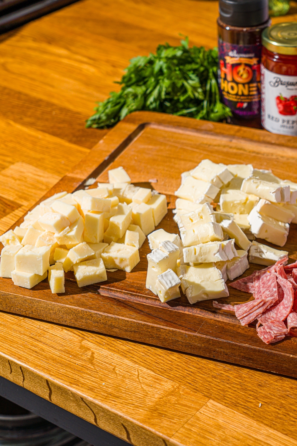 A cutting board with cubed cheddar cheese, brie, and additional meats. The board is on a wooden counter with fresh basil.