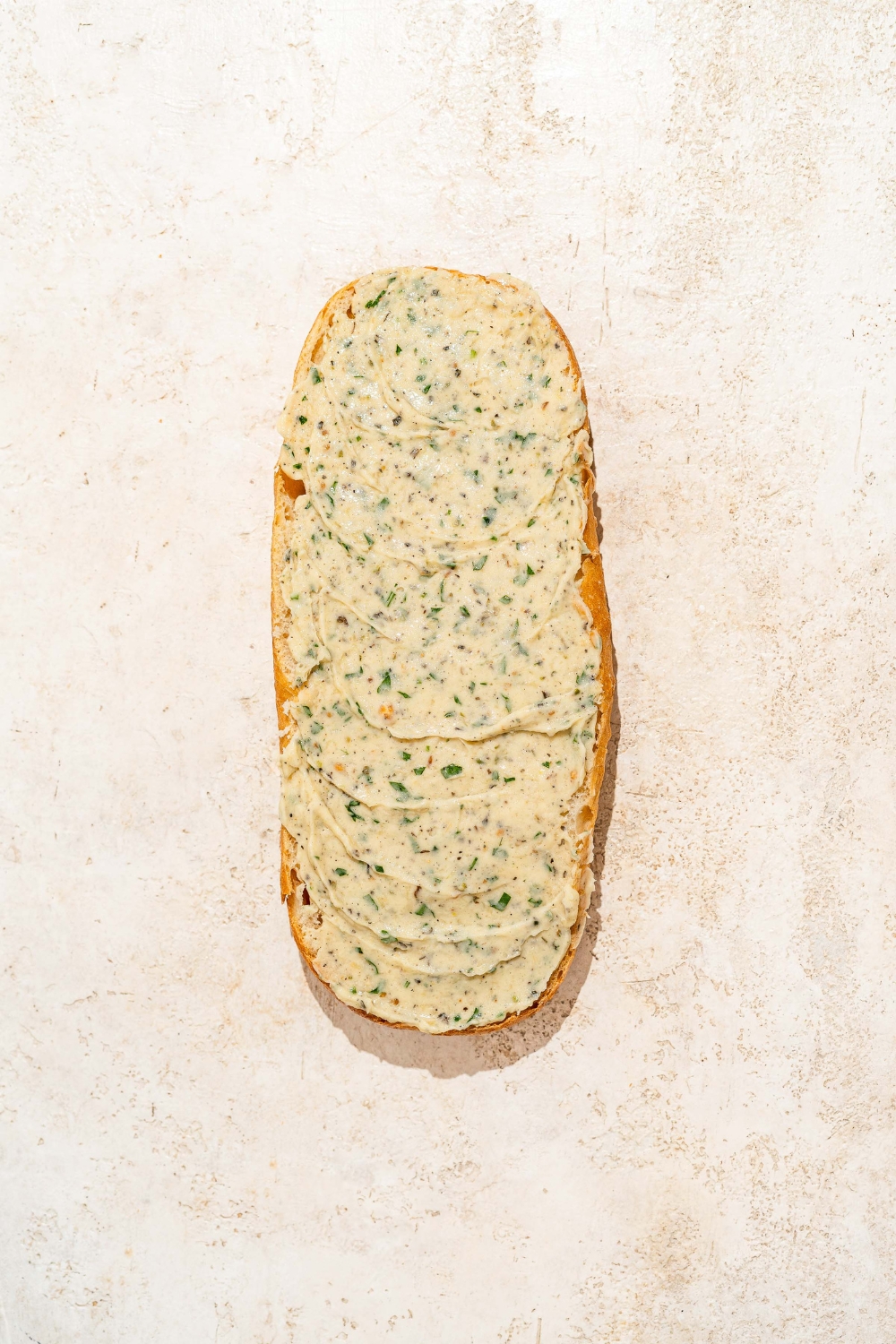 A cut side of an Italian bread loaf with garlic ranch butter spread on the bread. The bread is on a tan counter.