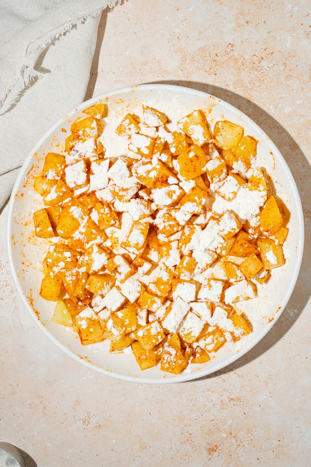 A white plate with seasoned cubed potatoes topped with flour and cornstarch. The plate is on a tan counter with a white cloth napkin.