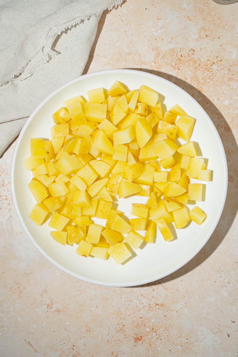 A white plate with cubed parboiled potatoes. The plate is on a tan counter with a white cloth napkin.