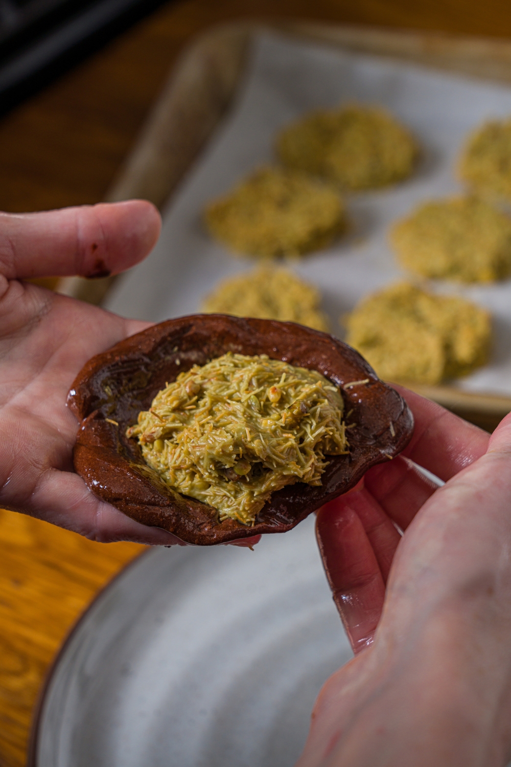 A close up of a hand wrapping Dubai chocolate around a pistachio filling ball. There is a baking sheet with pistachio fillings blurred in the background.