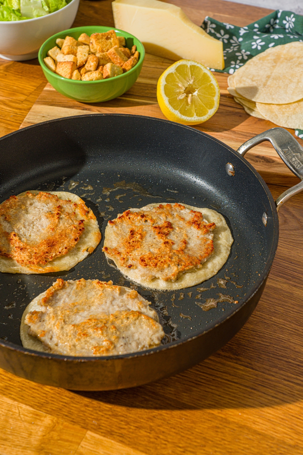 A skillet with three flipped chicken smash tacos cooking in oil. The skillet is on a wooden counter with a lemon and bowl of croutons.