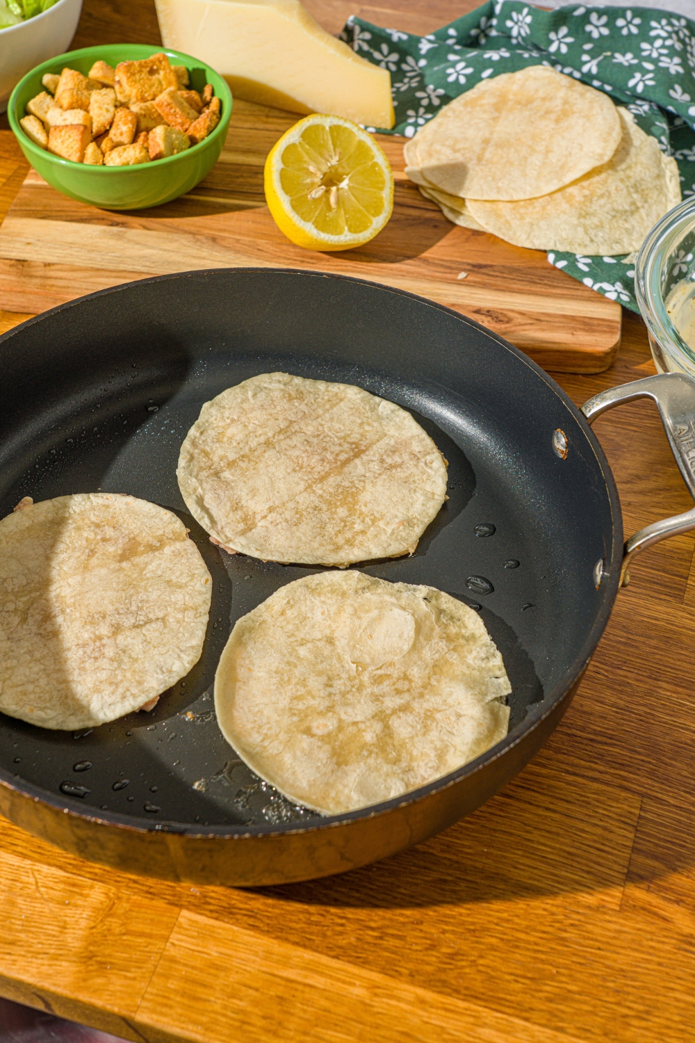 A skillet with three chicken smash tacos faced down. The skillet is on a wooden counter with a lemon and bowl of croutons.