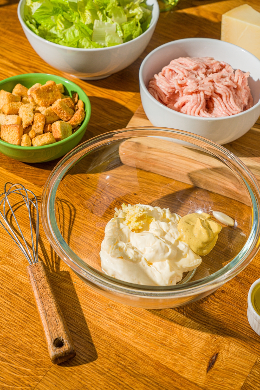 A glass bowl with ingredients to make caesar dressing including mayo, mustard, and garlic. The bowl is on a wooden counter with a whisk and bowls of ingredients.