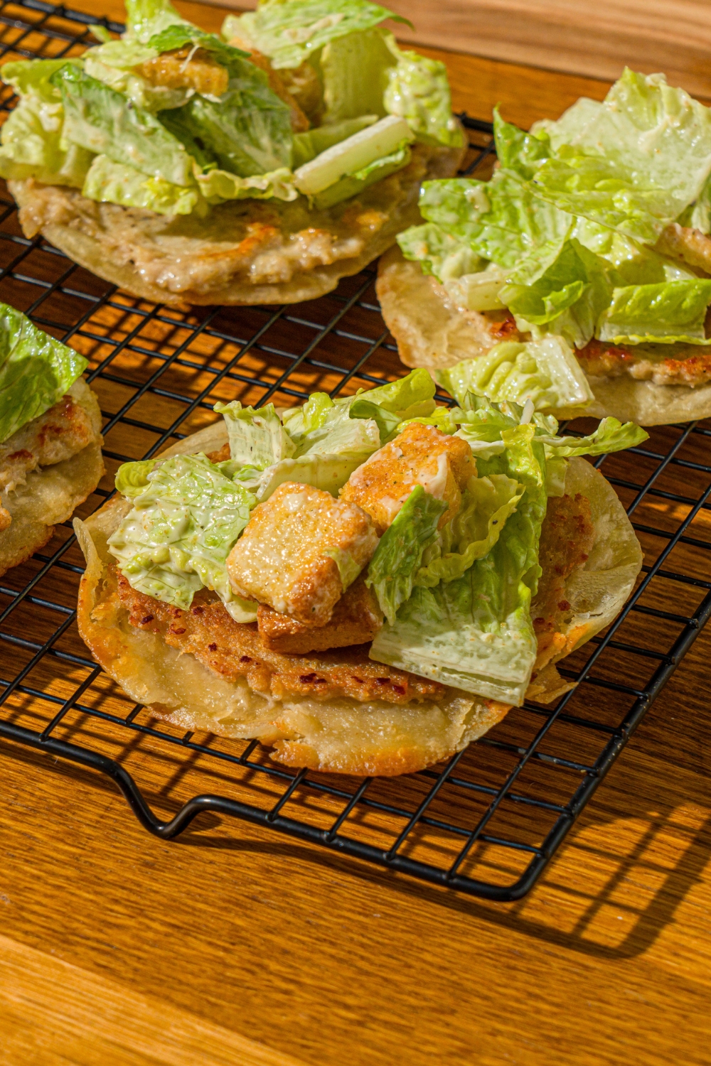 A wire rack with several open chicken caesar smash tacos. The rack is on a wooden counter.