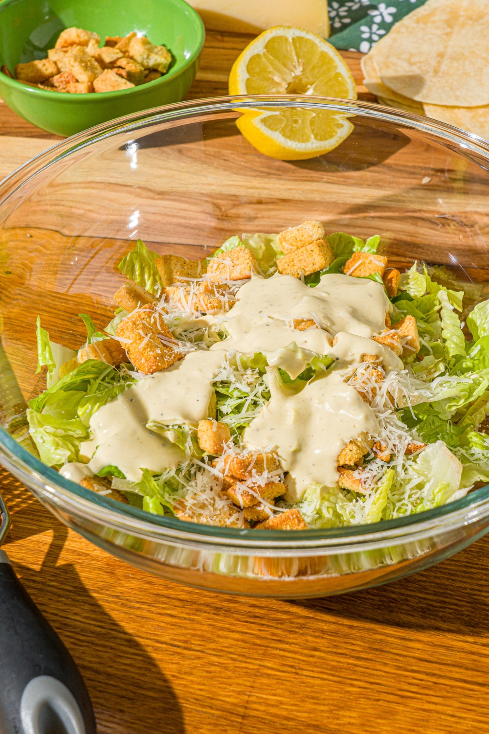 A glass bowl with romaine lettuce, shredded parmesan, and croutons topped with caesar dressing. The bowl is on a wooden counter with a lemon and bowl of croutons.