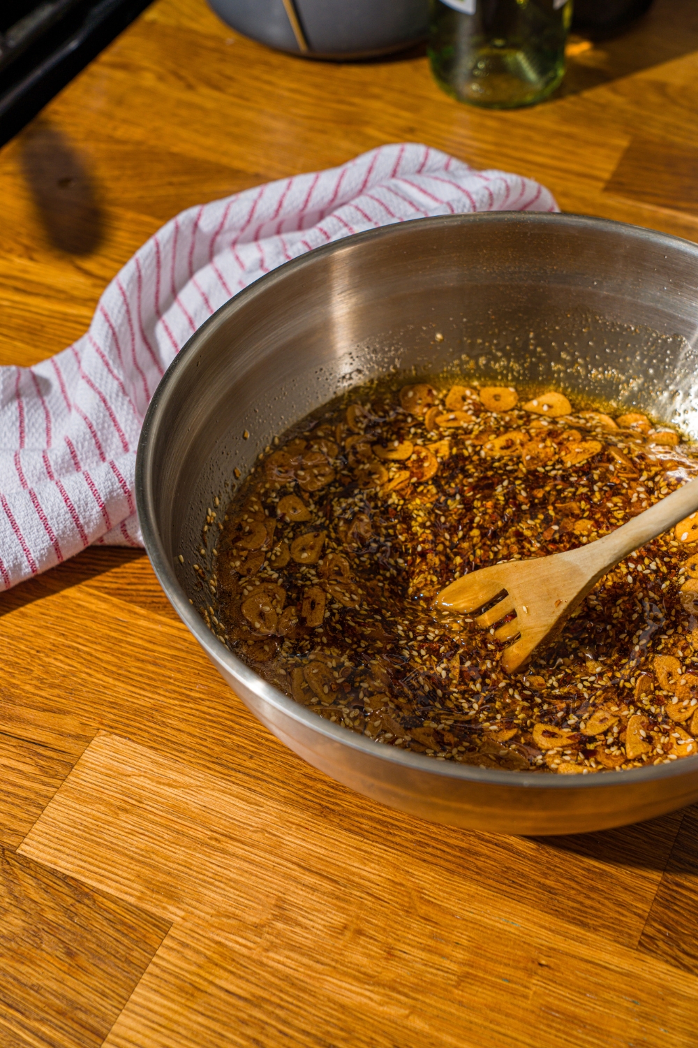 A large bowl with a wooden spoon stirring chili crisp oil mixture. The bowl is on a wooden counter with a white striped napkin.