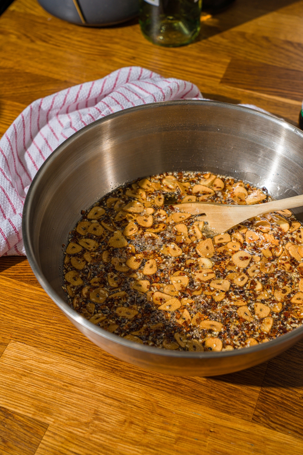 A large bowl with a wooden spoon stirring a chili crisp mixture. The bowl is on a wooden counter with a white striped napkin.